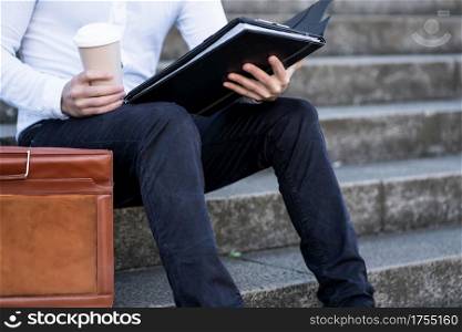 Businessman reading files and drinking a cup of coffee while sitting on stairs outdoors. Business concept.