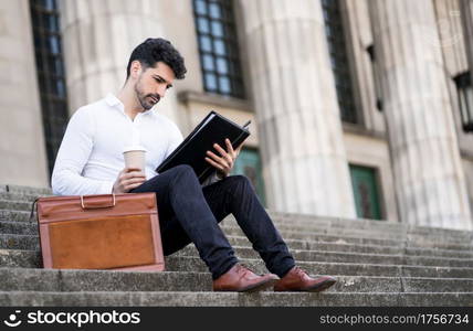 Businessman reading files and drinking a cup of coffee while sitting on stairs outdoors. Business concept.