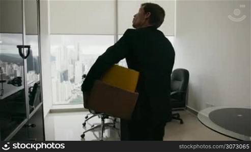 Businessman recently hired for corporate job moves into his new office. He holds a box with his folders and looks around with happy expression. Steadicam shot
