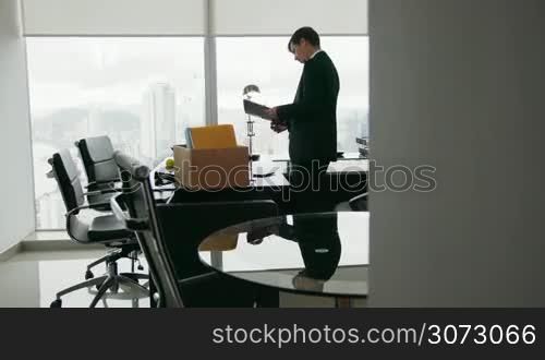 Businessman recently hired for corporate job moves into his new office. He takes out a folder and reads documents near a big window, contemplating the view of the city. Wide shot