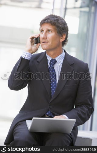 Businessman sitting in office lobby with laptop using cellular phone