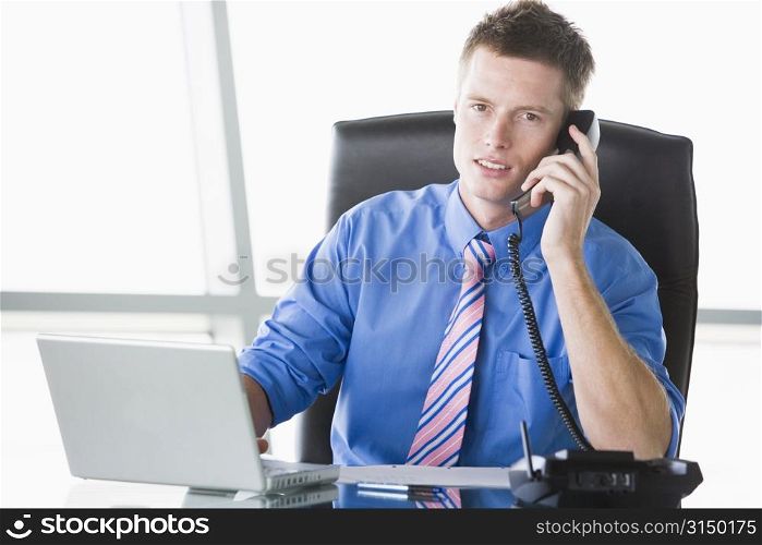 Businessman sitting in office with laptop using telephone
