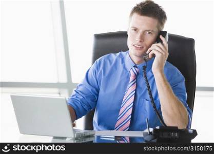 Businessman sitting in office with laptop using telephone