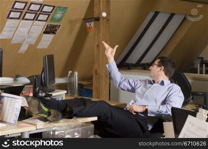 Businessman sitting on an office chair and throwing a crumpled paper ball in mini basketball hoop