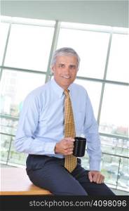 Businessman Sitting on Corner of His Desk With Coffee Mug
