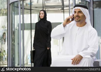 Businessman sitting outdoors by building using cellular phone with businesswoman walking in background (selective focus)