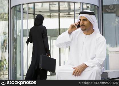 Businessman sitting outdoors by building using cellular phone with businesswoman walking in background (selective focus)