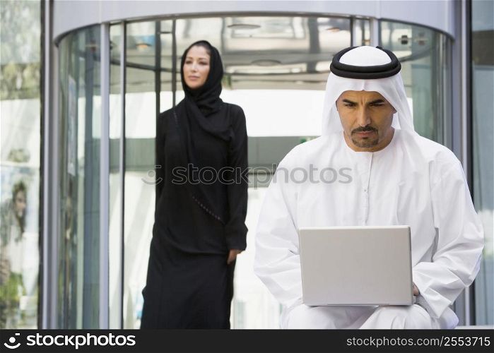 Businessman sitting outdoors by building using laptop with businesswoman walking in background (selective focus)