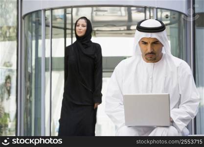 Businessman sitting outdoors by building using laptop with businesswoman walking in background (selective focus)