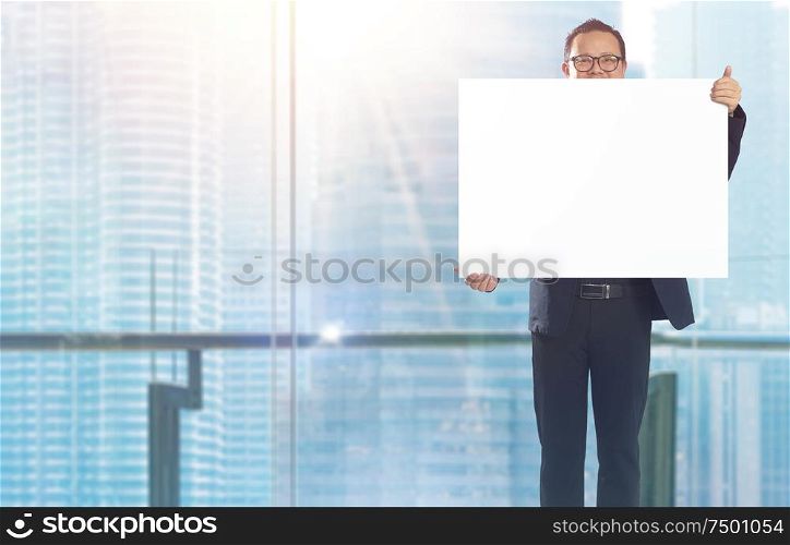 Businessman standing and holding white empty board in front of windows office overlooking the city at sunrise .