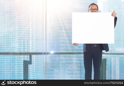 Businessman standing and holding white empty board in front of windows office overlooking the city at sunrise .