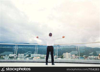Businessman standing keeping arms raised on a roof and looking at city Success concept