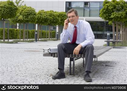 Businessman talking on mobile phone while sitting on a bench in front of an office building