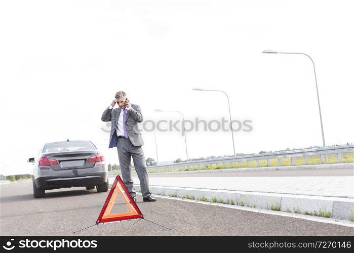 Businessman talking on phone while standing with breakdown car by warning sign on road