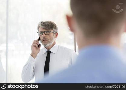 Businessman talking on smartphone with colleague by wall at office