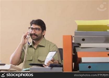 Businessman talking over telephone while looking at files sitting at his office desk.