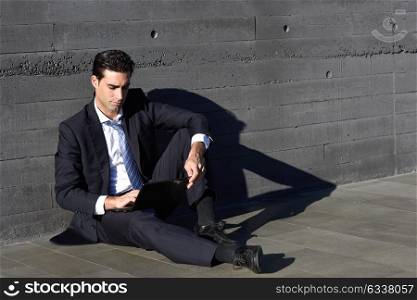 Businessman using a laptop computer sitting in the street. Man wearing blue suit and tie in urban background.