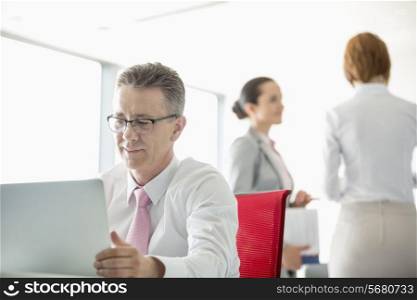Businessman using laptop with colleagues discussing in background at office