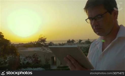 Businessman using touch pad against sunset on tropical resort, then he takes off glasses to rub his eyes