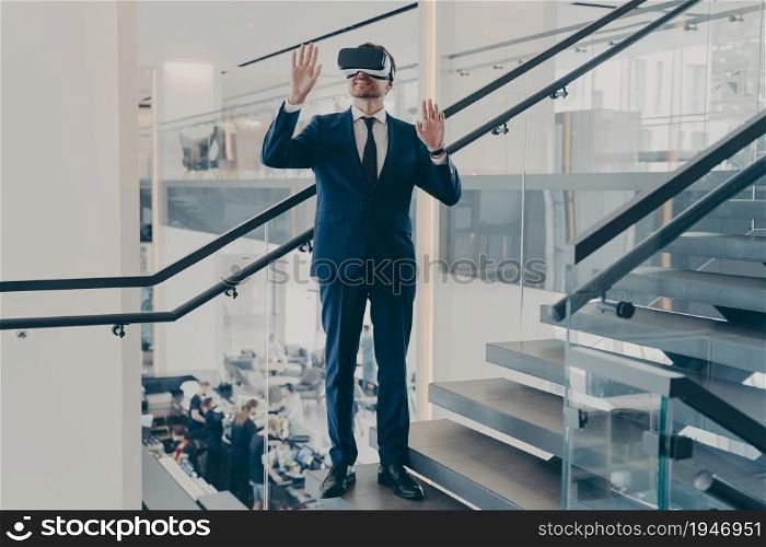 Businessman using virtual reality glasses in office lobby. Man in VR headset stands on staircase with hands up in air trying to touch objects in digital simulation. Innovative 3D technology concept. Businessman in VR headset stands on staircase in office lobby with hands up in air