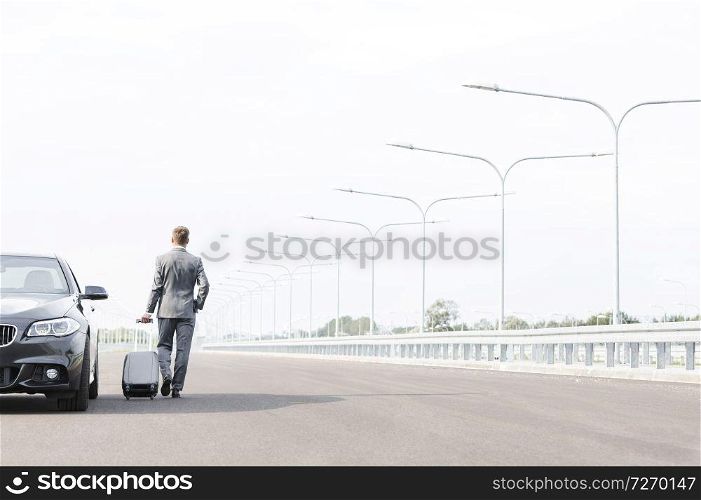 Businessman walking with suitcase by car on road against sky