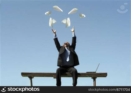 Businessman wearing black suit throws up all his papers in frustration as he sits on his park bench, next to his laptop