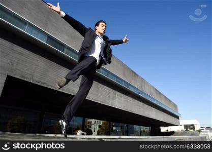 Businessman wearing blue suit and tie jumping with open arms in urban background. Man with formal clothes standing outside an office building.