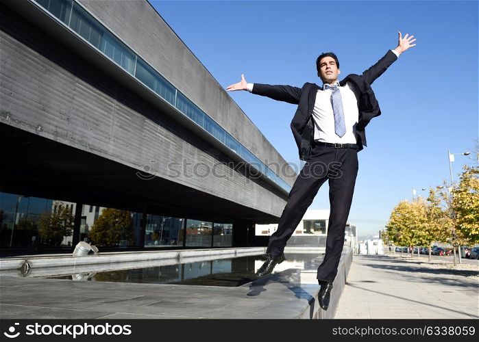 Businessman wearing blue suit and tie jumping with open arms in urban background. Man with formal clothes standing outside an office building.