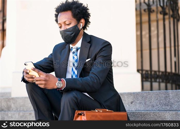 Businessman wearing face mask and using his mobile phone while sitting on stairs outdoors. New normal lifestyle. Business concept.