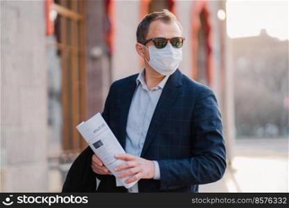 Businessman wears protective mask against transmissible infectious disease, reads newspapers, dressed elegantly, poses outdoor, thinks how to prevent new coronavirus from China. Flu in city.