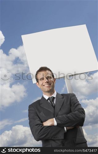 Businessman with arms crossed standing by blank sign against cloudy sky