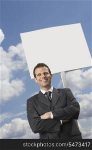 Businessman with arms crossed standing by blank sign against cloudy sky
