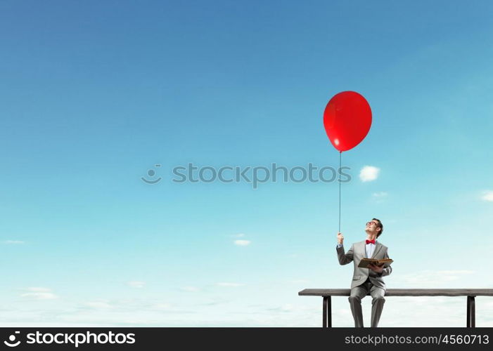 Businessman with balloon. Young businessman sitting on bench with balloon in one hand and book in other