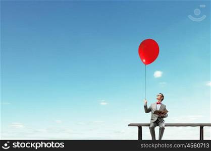 Businessman with balloon. Young businessman sitting on bench with balloon in one hand and book in other