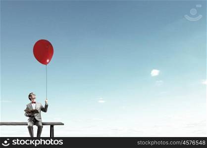 Businessman with balloon. Young businessman sitting on bench with balloon in one hand and book in other