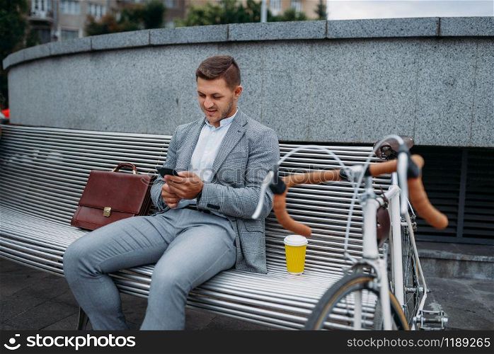 Businessman with bike talking by phone on the bench at the office building in downtown. Business person riding on eco transport on city street, urban style. Businessman with bike talking by phone on bench