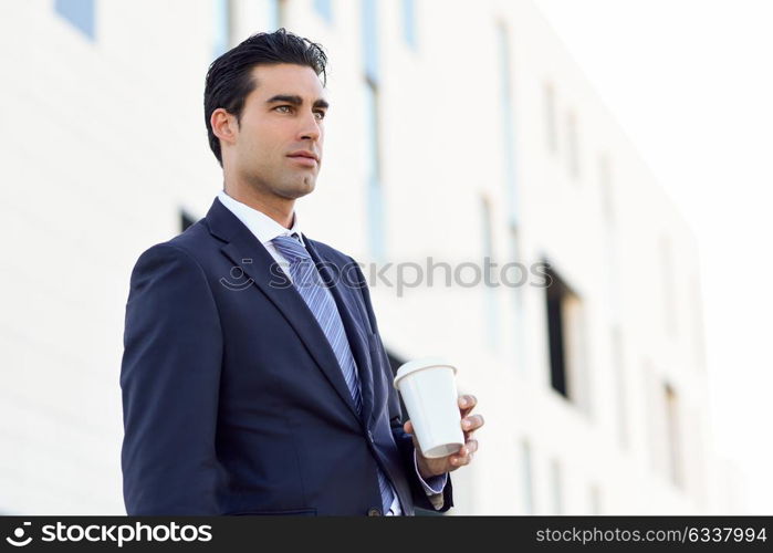 Businessman with formal clothes drinking coffee to go with a take away cup. Man wearing blue suit and tie in urban background.