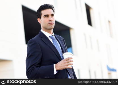 Businessman with formal clothes drinking coffee to go with a take away cup. Man wearing blue suit and tie in urban background.