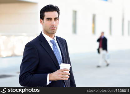 Businessman with formal clothes drinking coffee to go with a take away cup. Man wearing blue suit and tie in urban background.