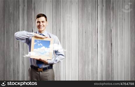Businessman with frame. Young smiling businessman holding wooden frame with sky picture