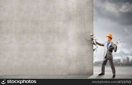 Businessman with hammer. Young businessman in suit and hardhat hammering nail in wall