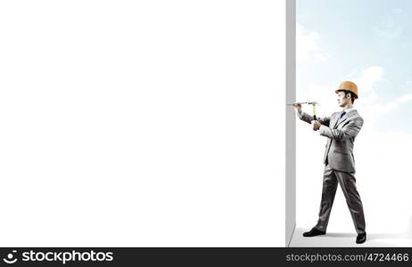 Businessman with hammer. Young businessman in suit and hardhat hammering nail in wall