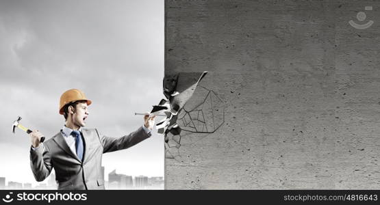 Businessman with hammer. Young businessman in suit and hardhat hammering nail in wall