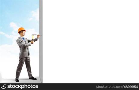 Businessman with hammer. Young businessman in suit and hardhat hammering nail in wall
