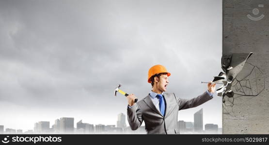 Businessman with hammer. Young businessman in suit and hardhat hammering nail in wall