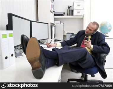 Businessman with his shoes on the desk, talking on the phone whilst making notes and holding another call