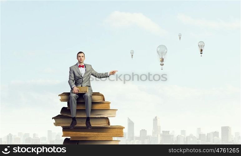 Businessman with old book. Young businessman sitting on pile of books and pointing with finger