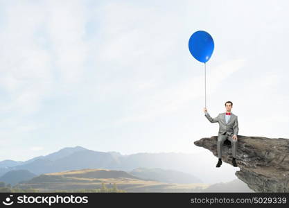 Businessman with red balloon. Young businessman wearing red bow tie sitting on rock edge with balloon