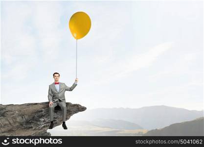 Businessman with red balloon. Young businessman wearing red bow tie sitting on rock edge with balloon