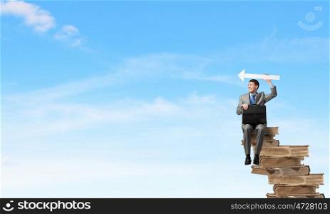 Businessman with signboard. Young businessman with suitcase sitting on pile of old books and showing direction